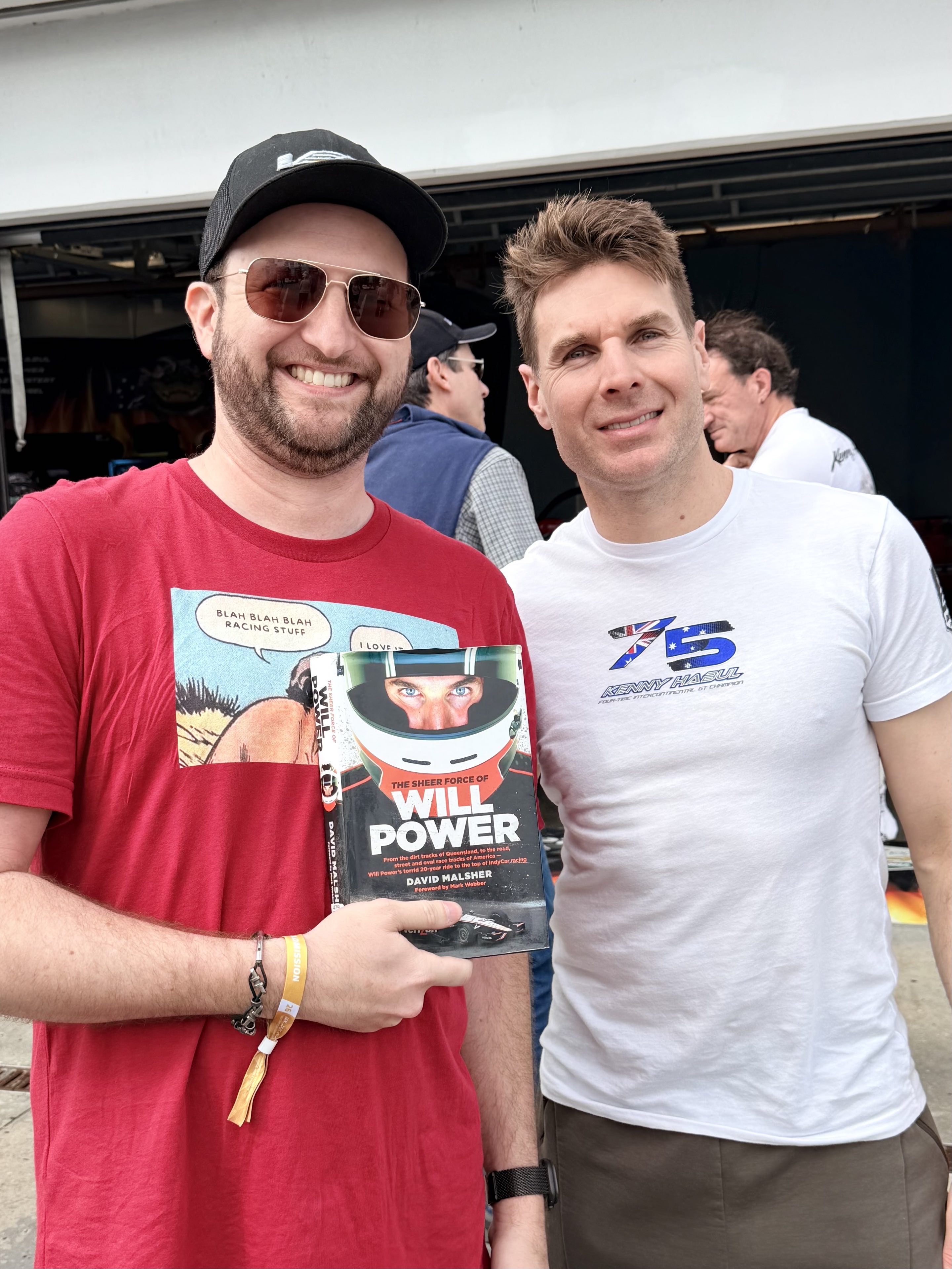Two men standing together in front of a race car garage, one holding a book that says Will Power on the cover, the other being Will Power